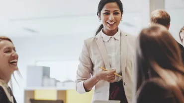 A woman in a suit smiling and talking to colleagues around a table in an office setting.