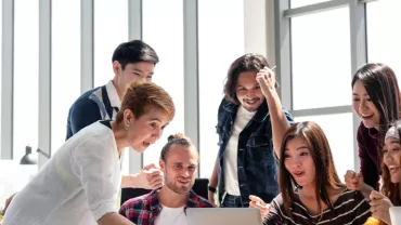 A group of people gathered around a laptop, looking surprised and delighted in a bright office setting.