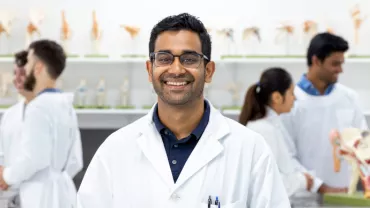 Smiling man in a lab coat stands in a scientific classroom with anatomical models in the background.