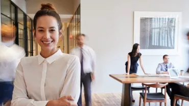 Smiling woman in white shirt stands with blurry office workers in background.