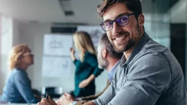 Smiling man with glasses in a meeting room with colleagues.