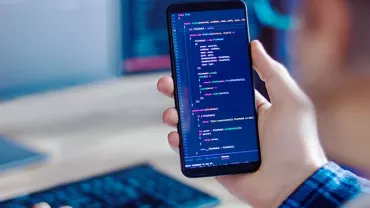 Person holding smartphone with code on screen, working at a computer.