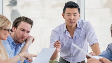 A diverse group of people in a meeting, discussing documents around a table.