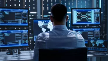 Person in front of multiple monitors displaying data and maps in a control room.