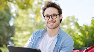 Person with glasses smiling outdoors on a sunny day.