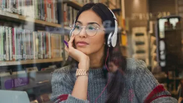 Woman with glasses and headphones, sitting in a library, looking thoughtful.