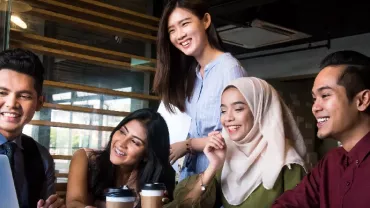 Five people smiling and looking at a laptop screen in a modern office setting.