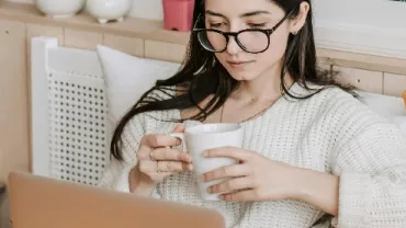 Woman wearing glasses, holding a mug, and using a laptop.