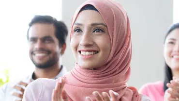 A smiling woman in a pink hijab applauds with others blurred in the background.