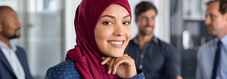 A female professional smiling, on a office background with three people dressed in corporate wear seated behind her